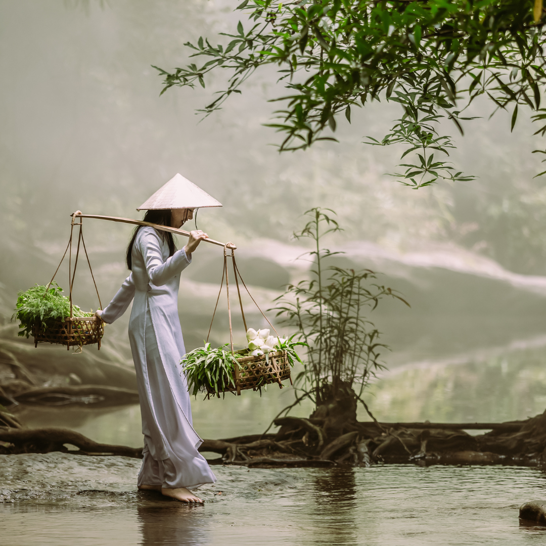 Woman in traditional Vietnamese Ao Dai and conical hat carrying baskets across a misty river
