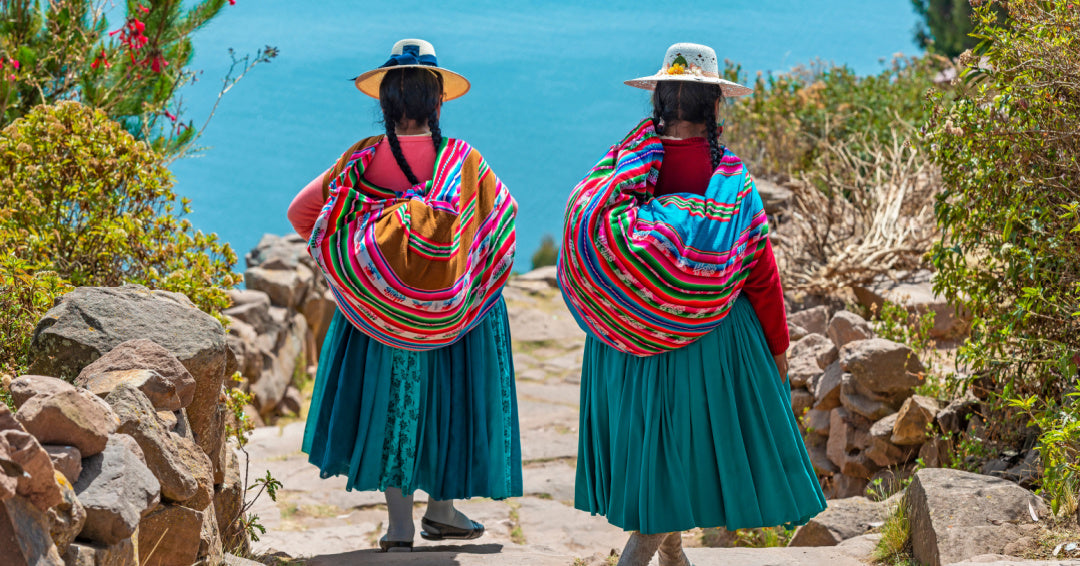 Two women in traditional Andean dress with colorful woven shawls walking by the sea.