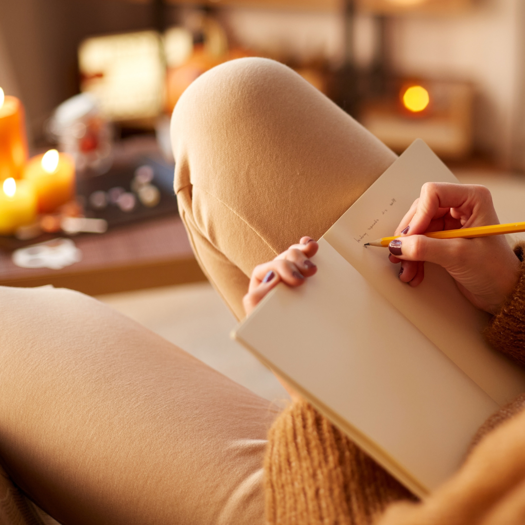 Person in a cozy brown sweater writing in a notebook by candlelight.