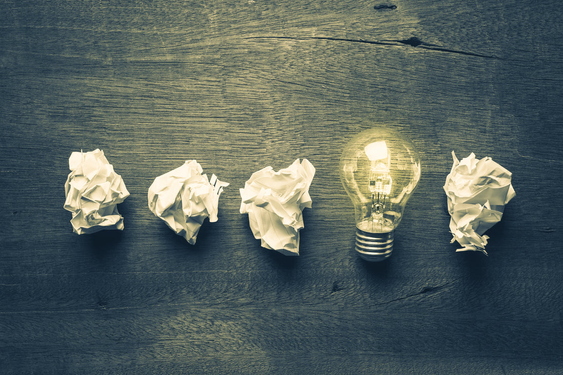 A glowing lightbulb centered among crumpled pieces of paper on a dark wooden surface.