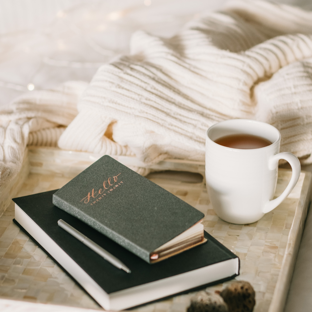 A cozy setup with a 'Hello' notebook, pen, and a cup of tea on a tray next to a white knit sweater.