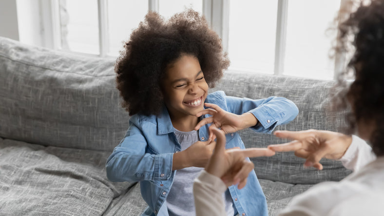 A joyful young girl with curly hair laughing while playing hand games with an adult on a sofa.