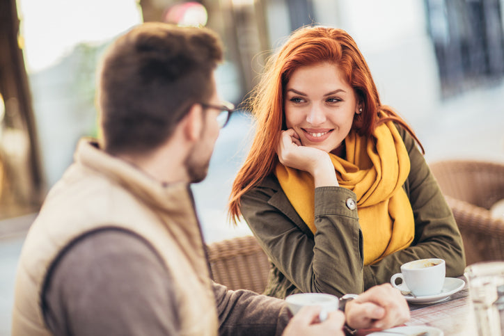 A red-haired woman in a yellow scarf smiling at a man in a cafe setting.