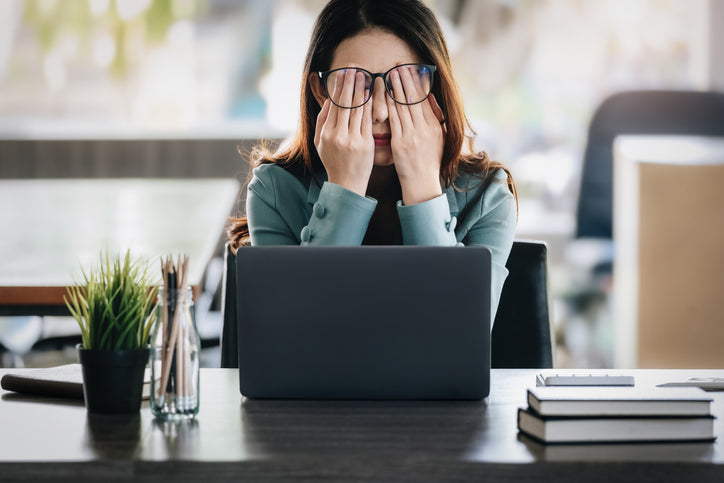 Stressed woman with glasses covering her face in front of a laptop at a desk.